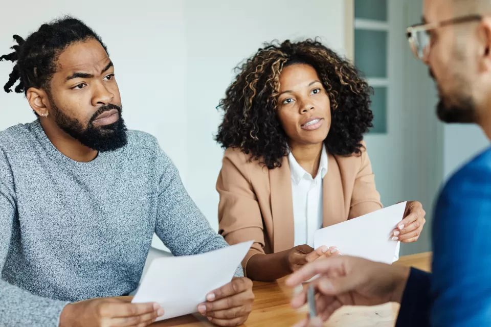 A Black couple holding contracts sits across the desk from a white mortgage lender.